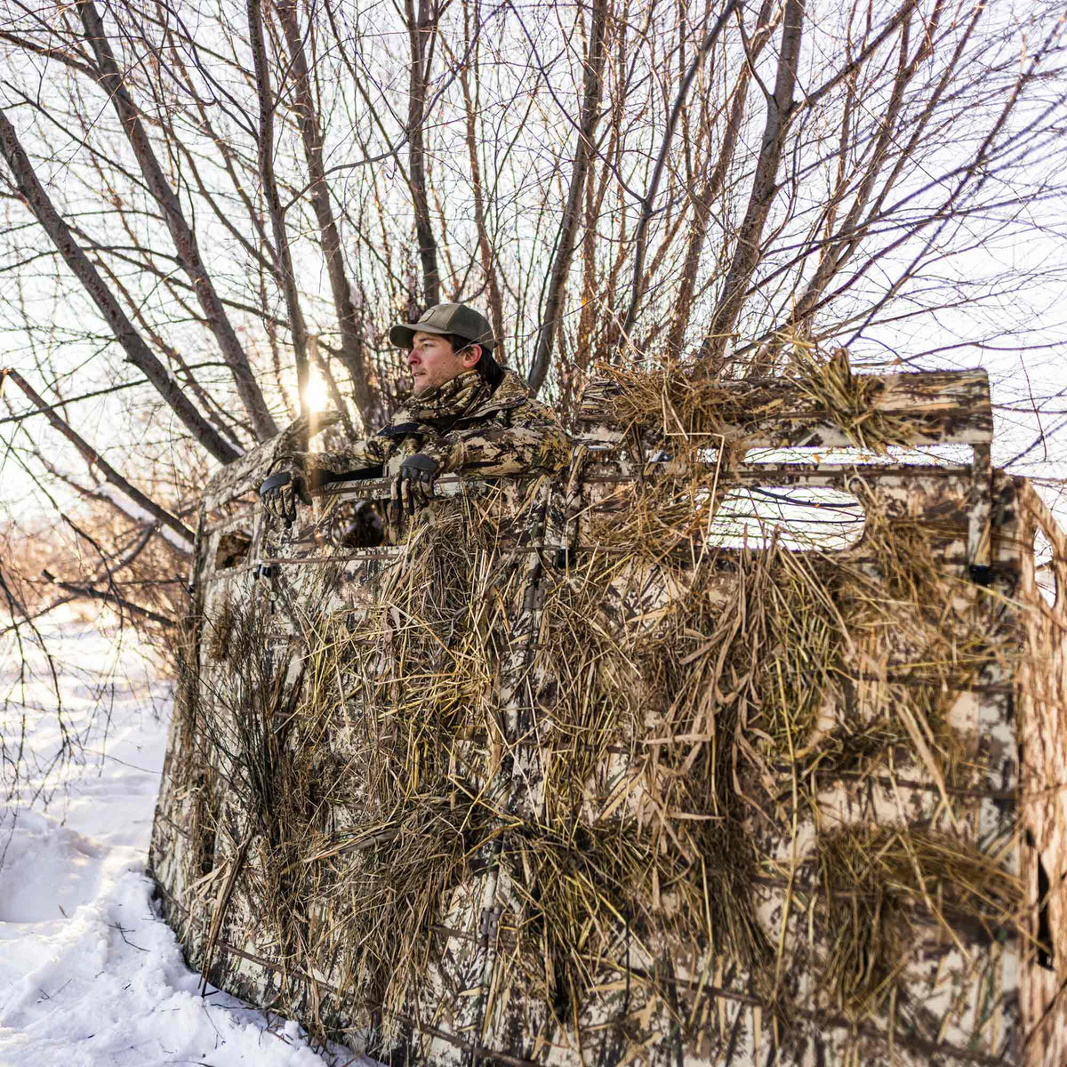 A man in camouflage sits in a hunting blind with a dry grass/reeds camouflage pattern, blending into the snowy, treed winter landscape.