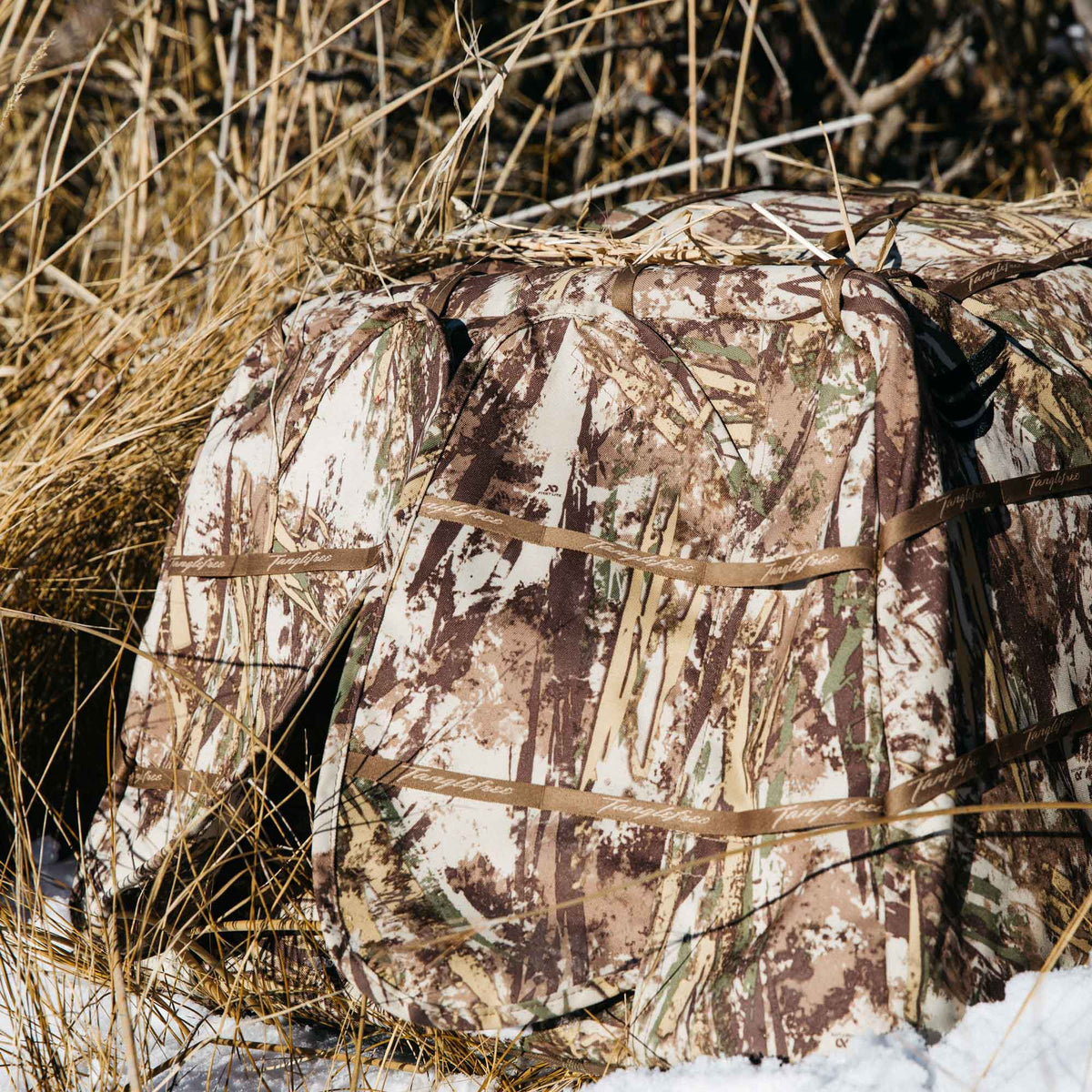 Multi-color camouflage hunting blind with Tanglefree branded straps, partially hidden in dry grass and snow.