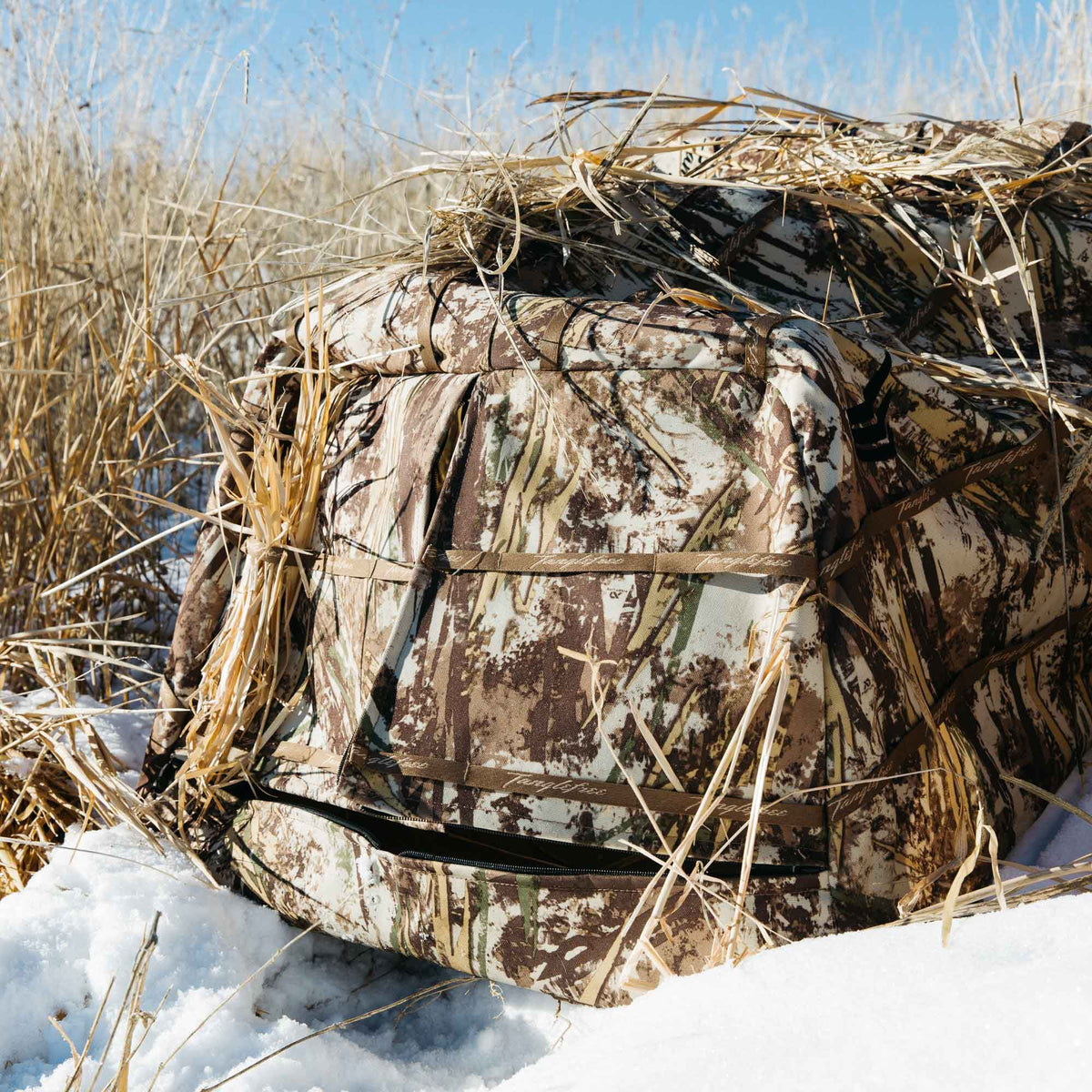 A camouflage hunting blind with vegetation straps and a partially unzipped opening, blending into dry grass and snow.