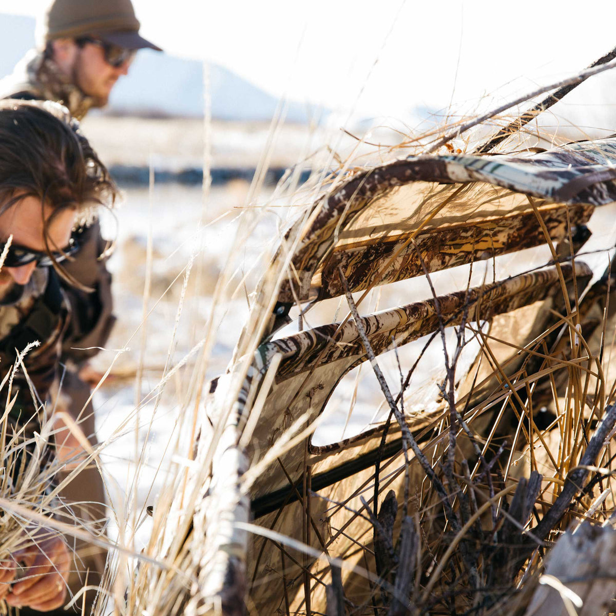 A camouflage field hunting blind blending into dry grass, with two blurred hunters in the sunny background.