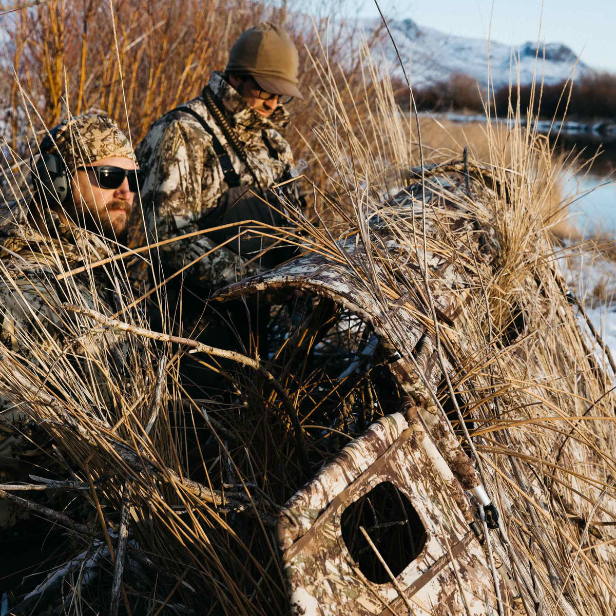 Camouflage hunting blind covered in dry reeds and grass, with an open window, used by two hunters.