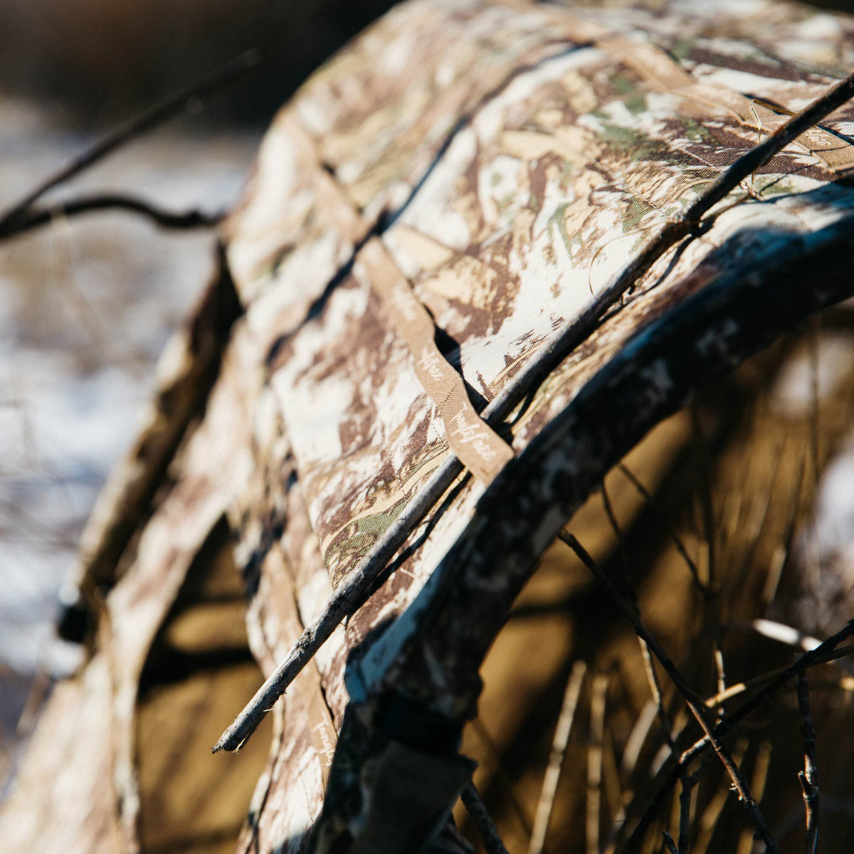Camouflage hunting blind with natural branches draped over it and a visible Tanglefree brand strap.
