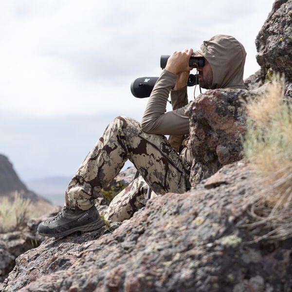 Man in tan hooded shirt, camo pants & dark gray SCHNEE hiking boots sits on rocks.