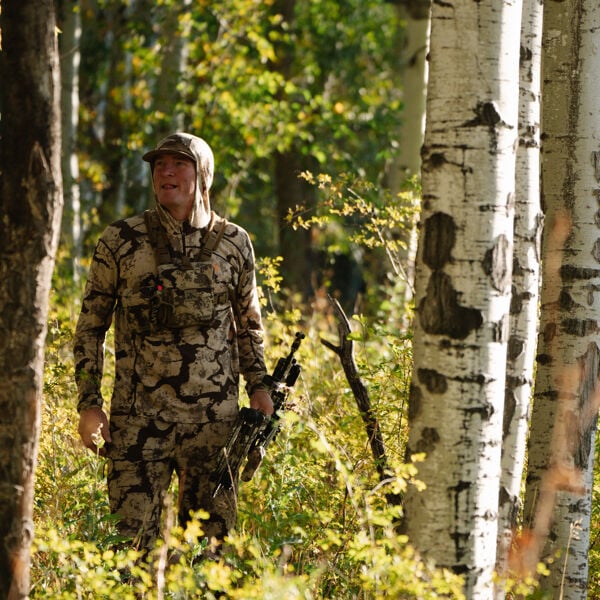Man in earthy camo hunting hoodie, pants, and chest rig, holding a bow in a forest.
