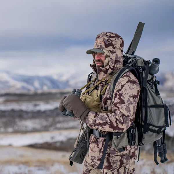Brown/tan camo hooded jacket and cap worn by hunter holding binoculars in snow.