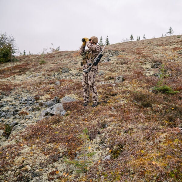 Hunter in brown camouflage suit scanning with binoculars, rifle on shoulder.
