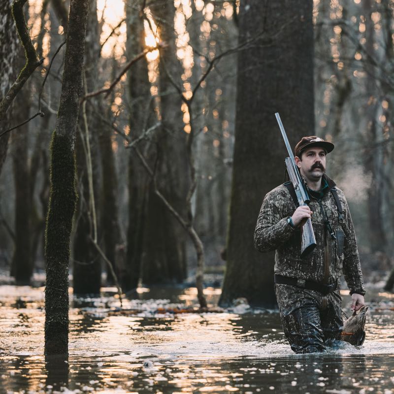Hunter in brown camo outfit wades in swamp, carrying shotgun and duck.