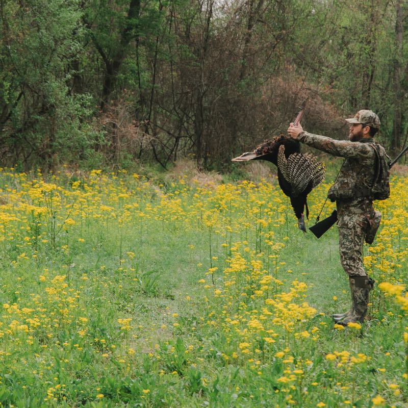 Hunter in camo holding a black turkey, shotgun slung, in a field of yellow flowers.
