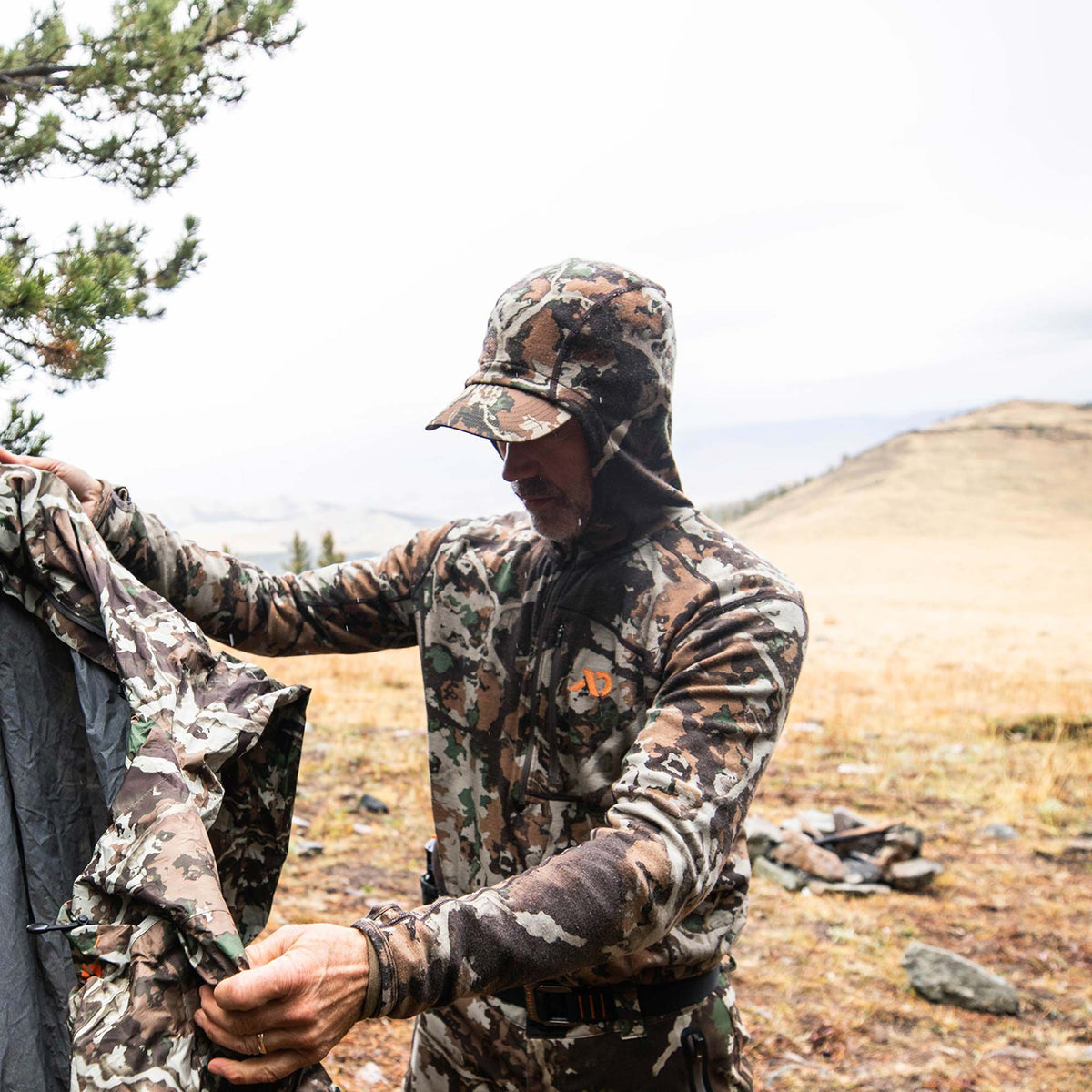 A man wears a camouflage hooded quarter-zip jacket with an orange chest logo and matching pants, holding a folded camouflage garment.