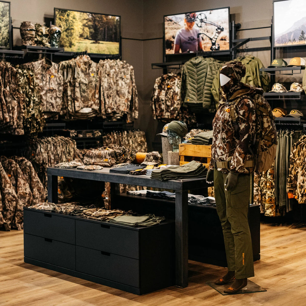 Mannequin in First Lite camo hunting jacket, pants, & backpack, with more apparel nearby.