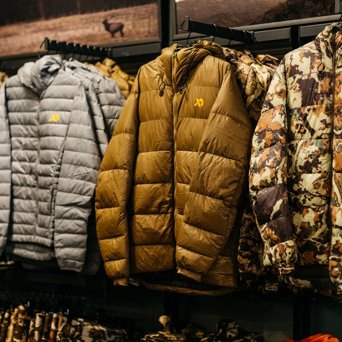 Row of grey, brown, and camo puffy jackets with yellow logos on a display rack.
