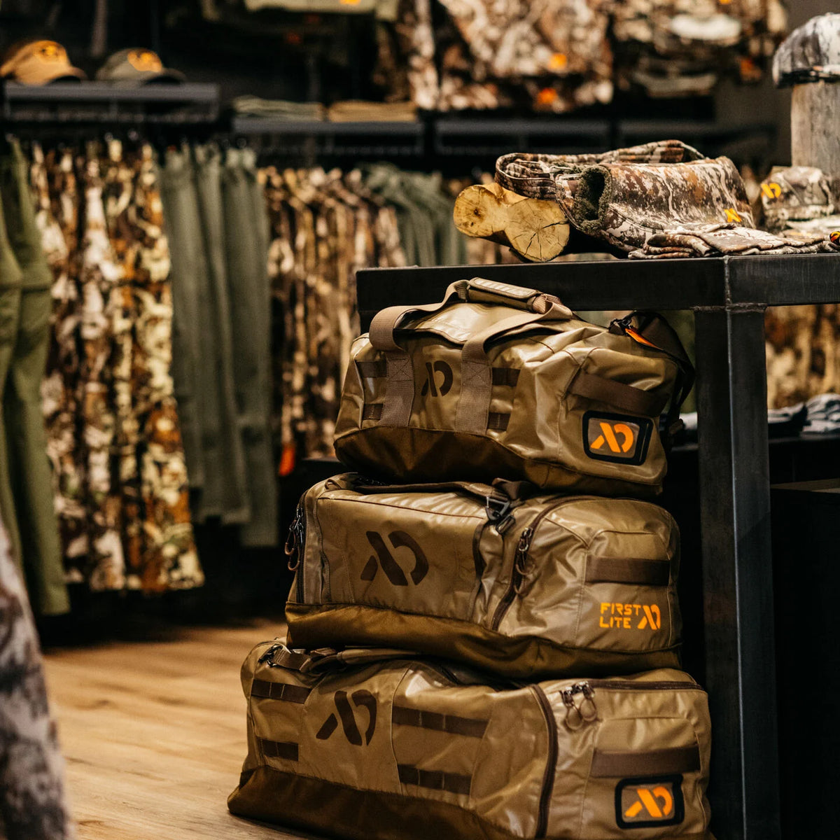 Three stacked tan First Lite duffel bags with orange logos.