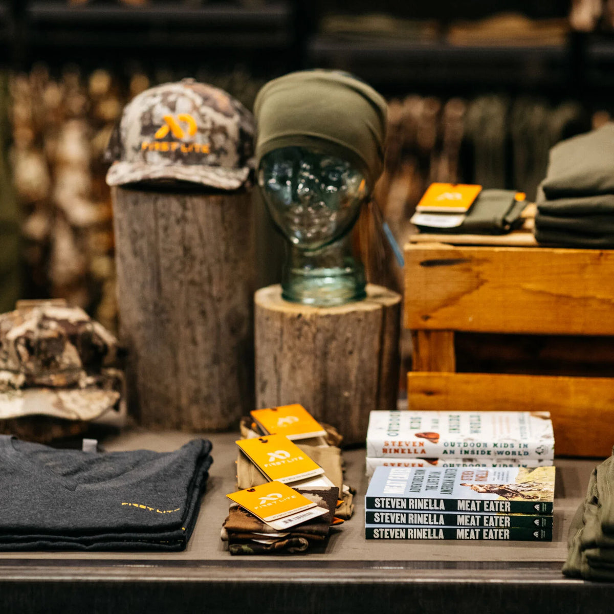 First Lite camo hat, olive beanie, charcoal shirt, and Steven Rinella's MeatEater books displayed.