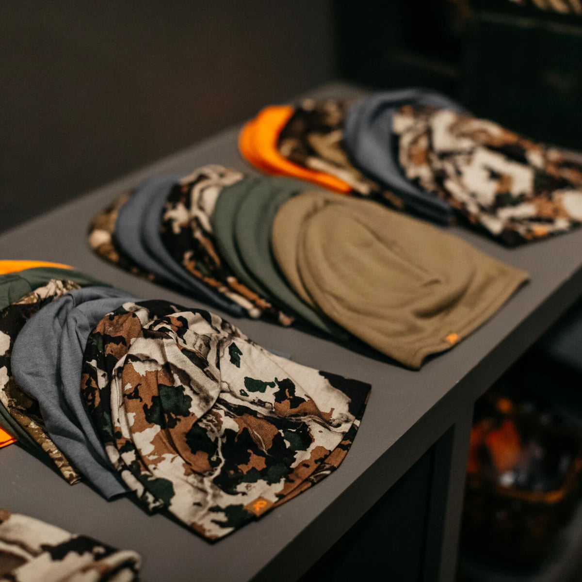 Stacked beanies in various camo patterns, gray, green, tan, and orange on a shelf.