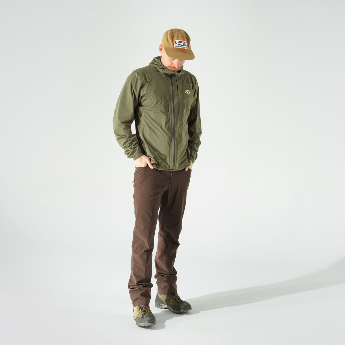 A man wears an olive green hooded zip-up jacket, brown pants, and a tan baseball cap, standing with hands in pockets.