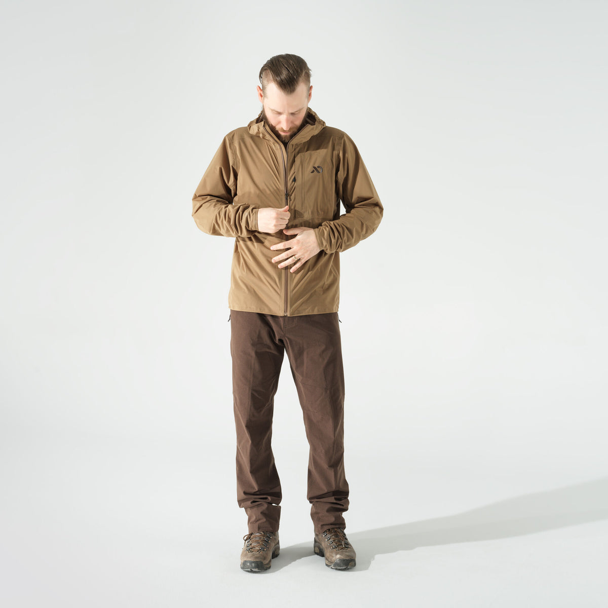 Man zipping up a tan hooded jacket with a dark logo on the left chest.