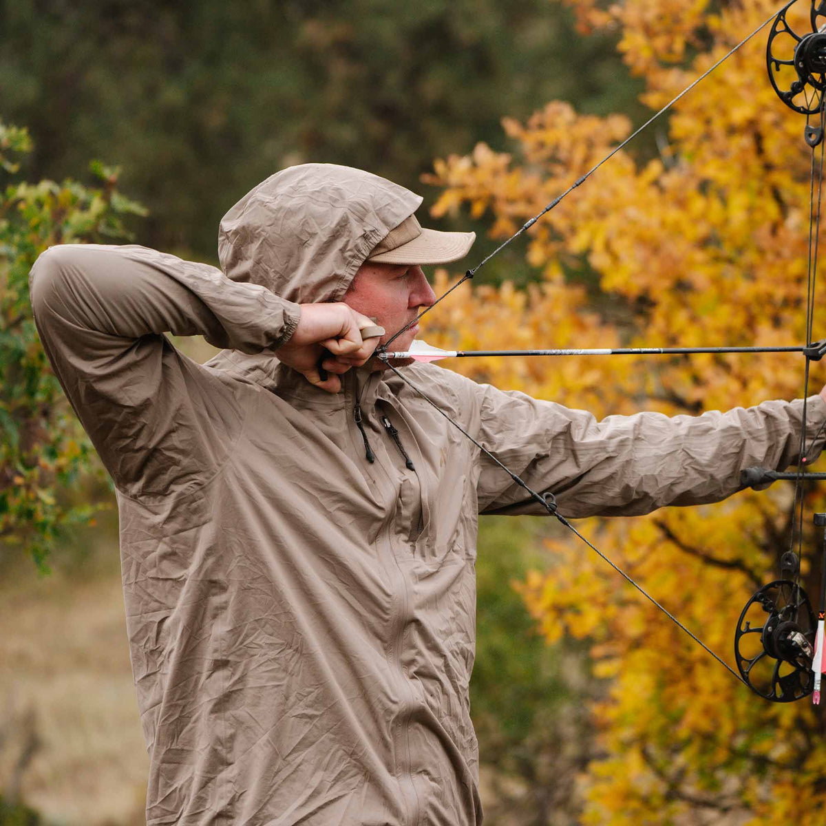A person in a tan lightweight hooded jacket with drawstrings aiming a bow and arrow.