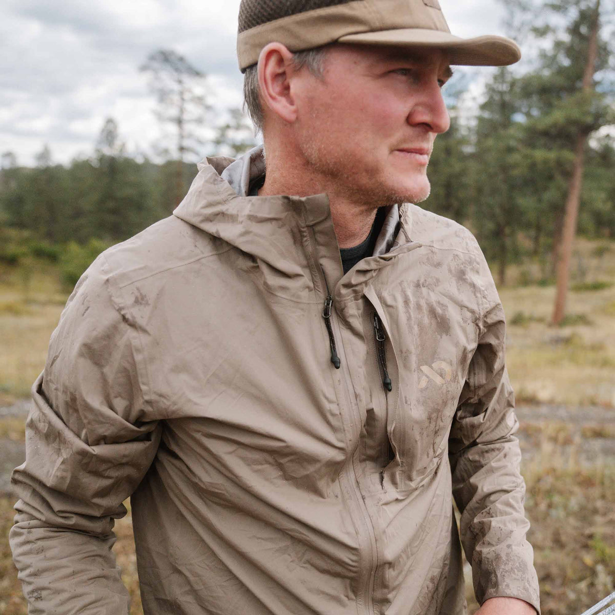 Man wearing a dirt-stained tan hooded jacket with a zippered chest pocket outdoors.