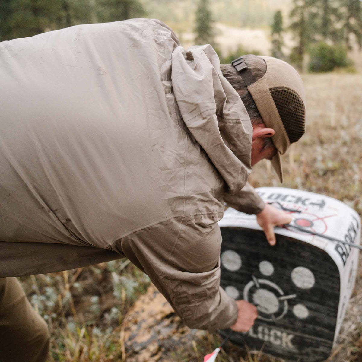 A person wears a tan hooded lightweight jacket with visible dirt marks while bending over an archery target.