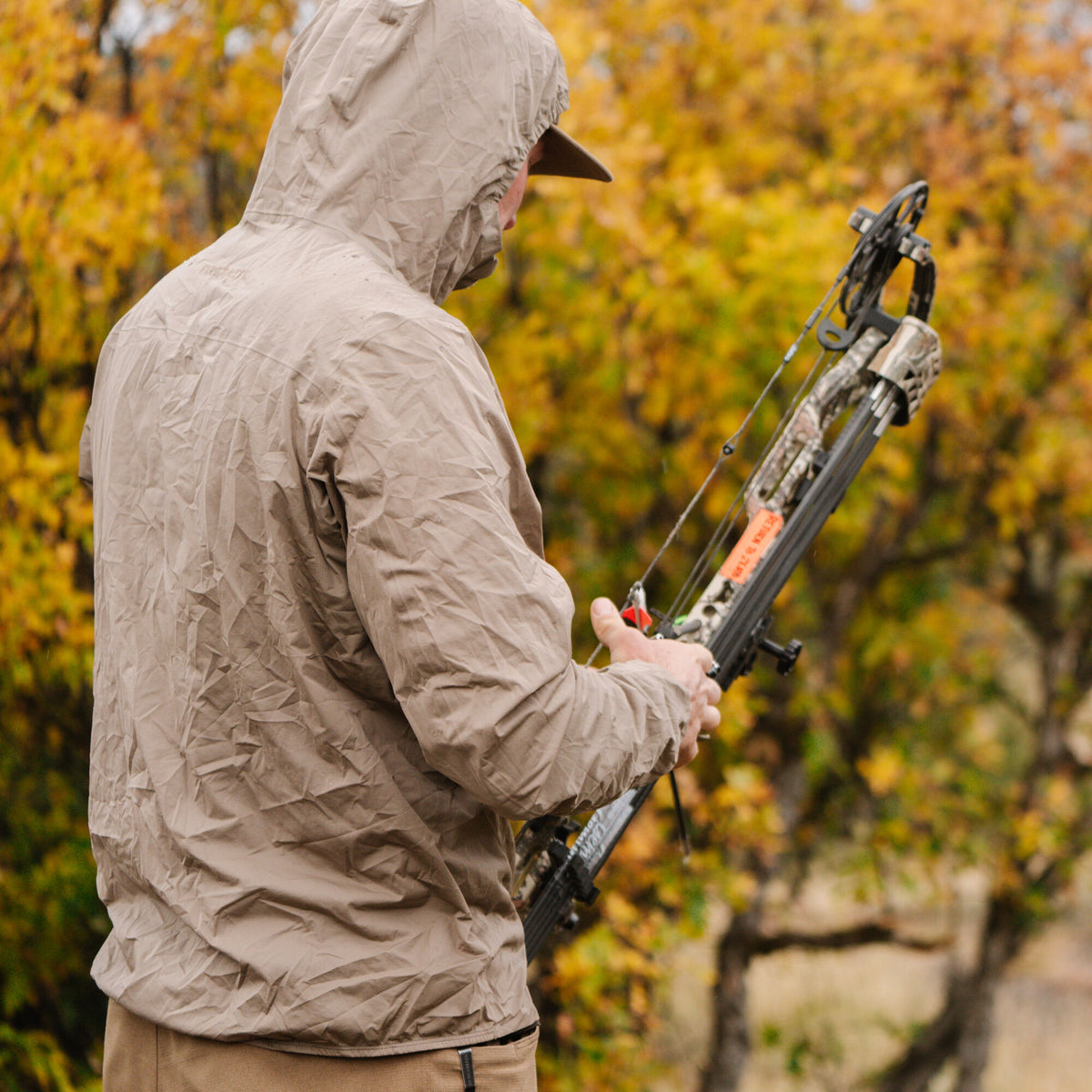 A person wears a tan crinkled rain jacket with the hood up, holding a camouflage compound bow.