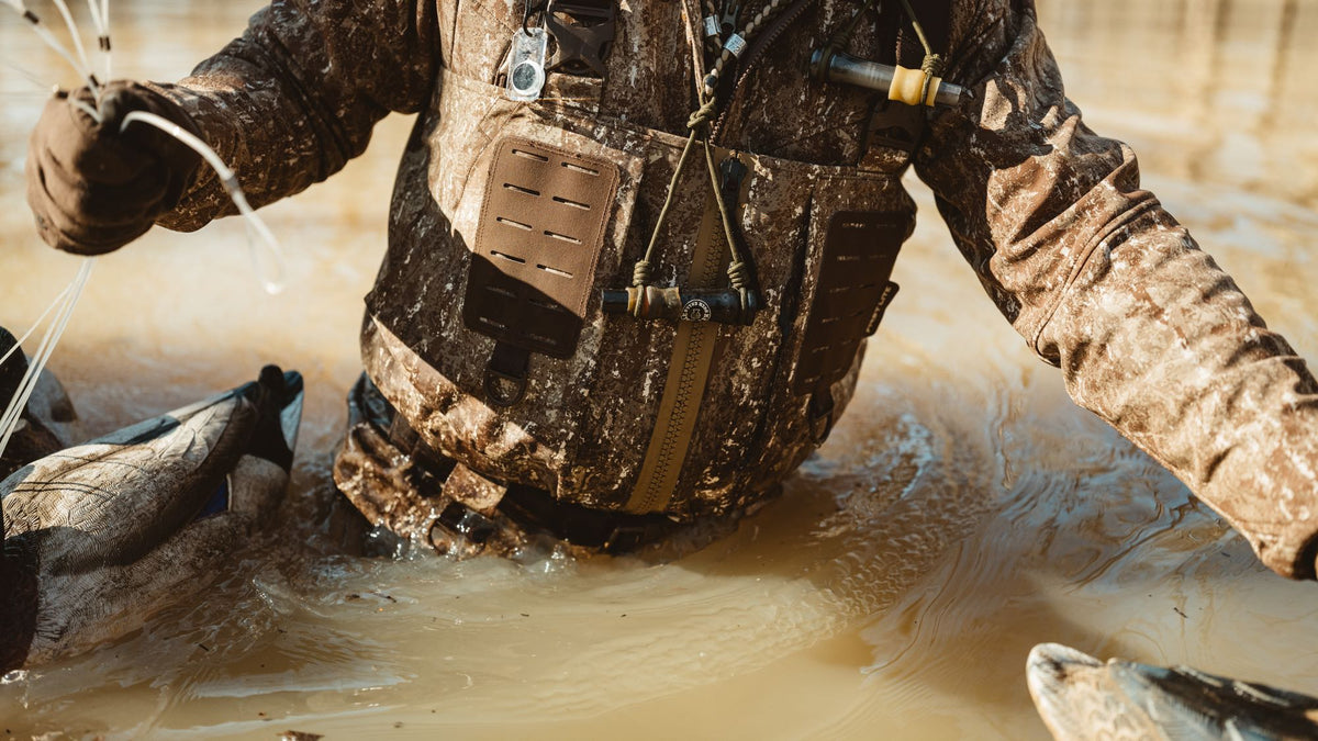 Brown marsh camo waders with duck call and gear loops, worn by hunter in muddy water.
