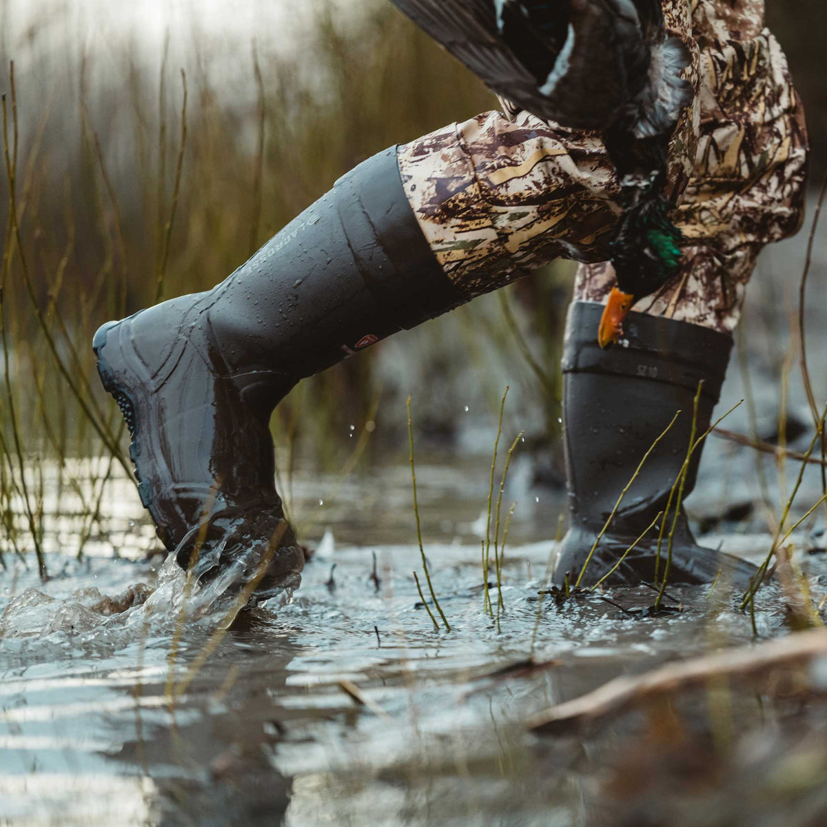 Dark grey LaCrosse hunting boots worn by a person in camouflage pants wading through splashing water while carrying a duck.