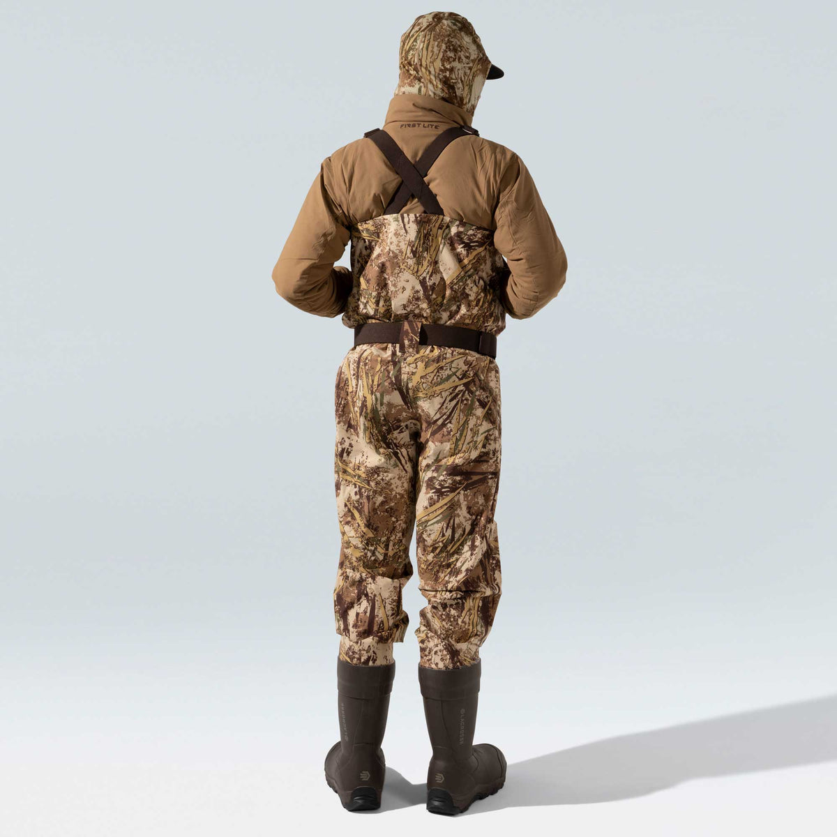 Rear view of a person wearing First Lite camouflage waders with dark suspenders and belt, a tan jacket, and dark brown LaCrosse boots.
