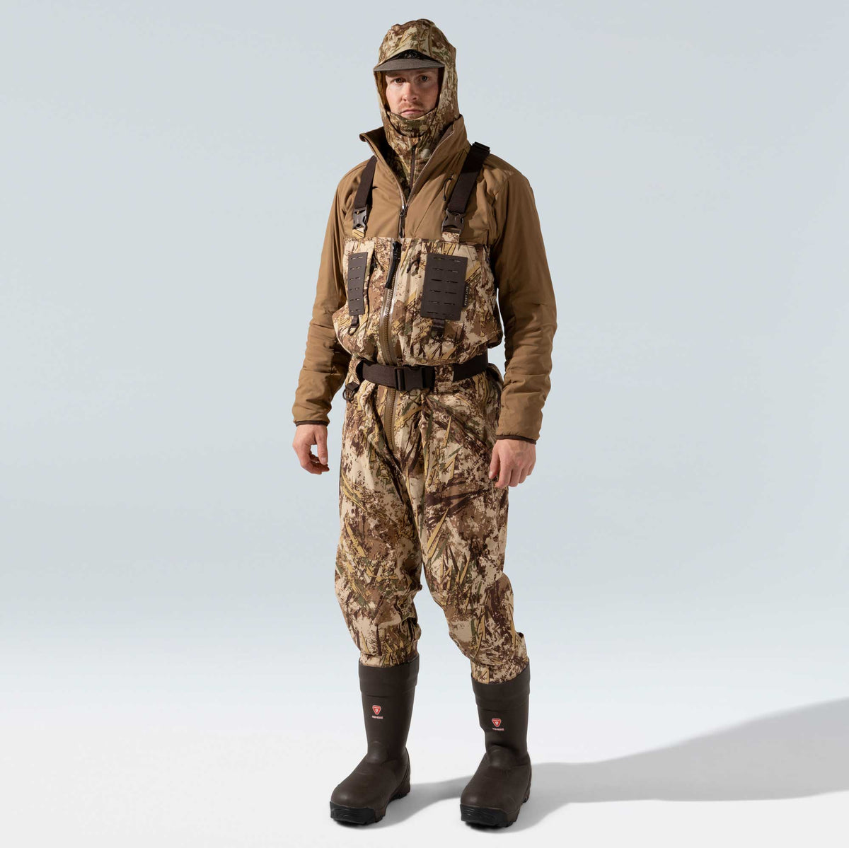 Man wears a brown hooded jacket over camouflage waders with suspenders, a belt, and integrated dark brown boots.