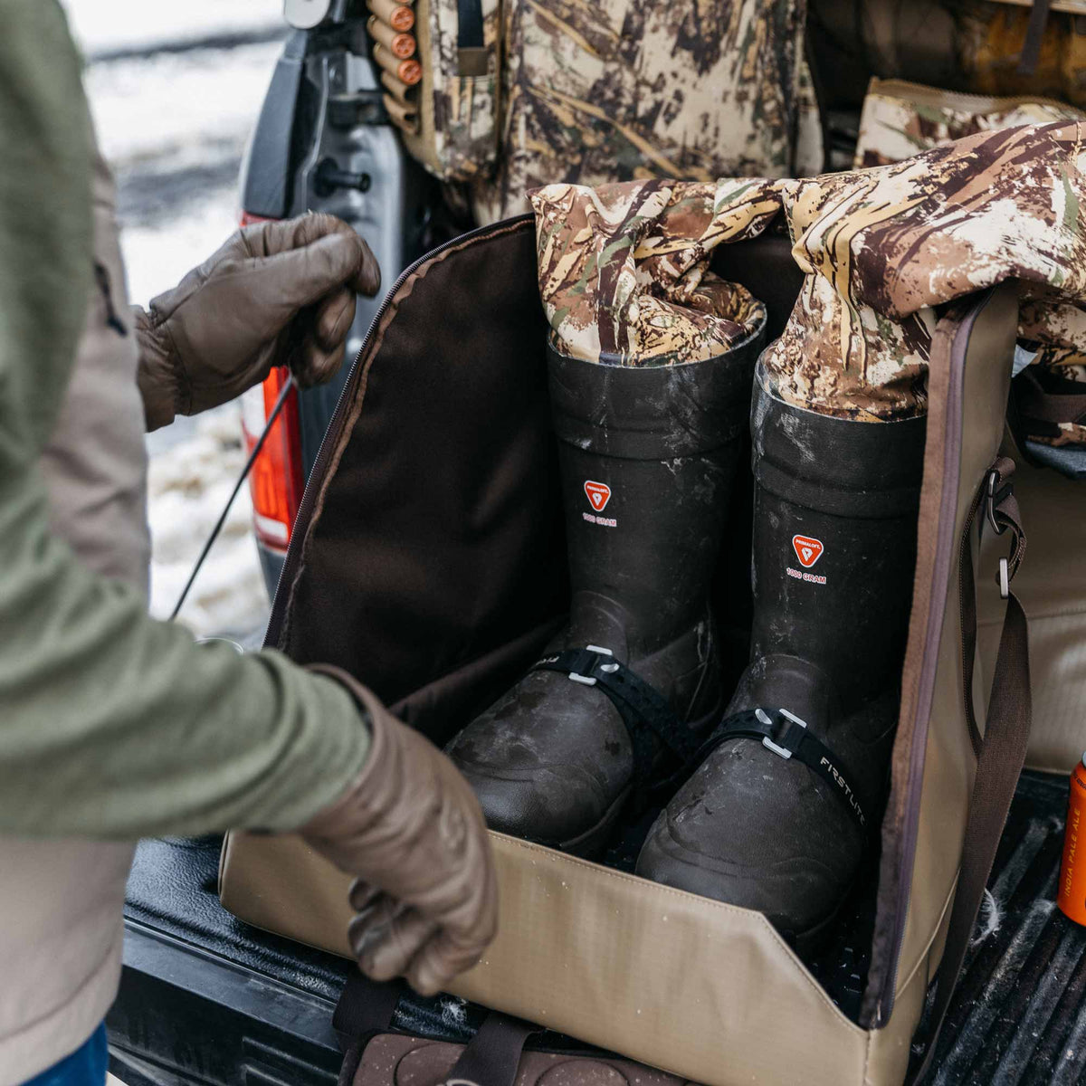 Black rubber hunting boots with camouflage fabric uppers and "Firstlite" straps, stored in a tan boot bag on a truck bed, with a person's gloved hands.