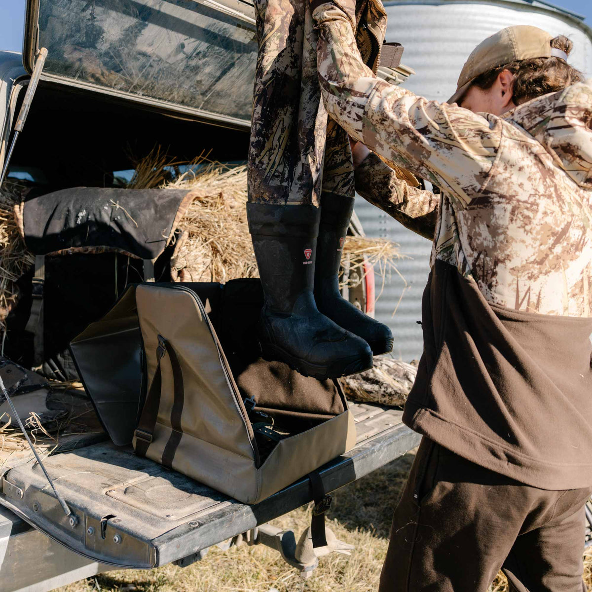 Camouflage waders with black rubber boots featuring a red shield logo are held by a person in a truck bed.