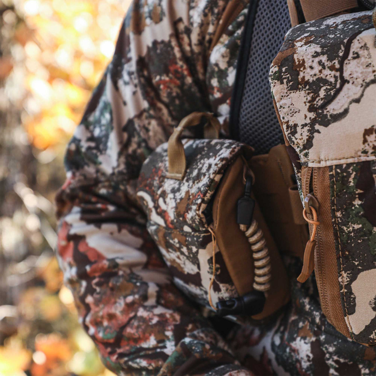 Camouflage bino harness with tan accents, zippered pouches, and a coiled tether worn over camouflage clothing.