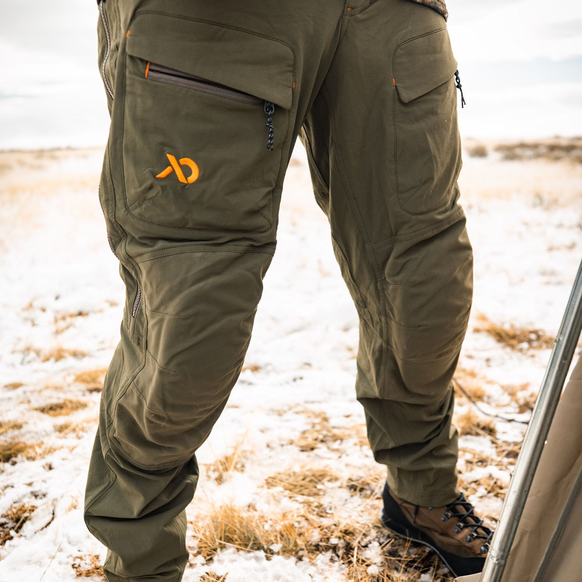 Olive green outdoor pants with an orange logo, multiple thigh pockets including zippered and flap styles, and reinforced knees.