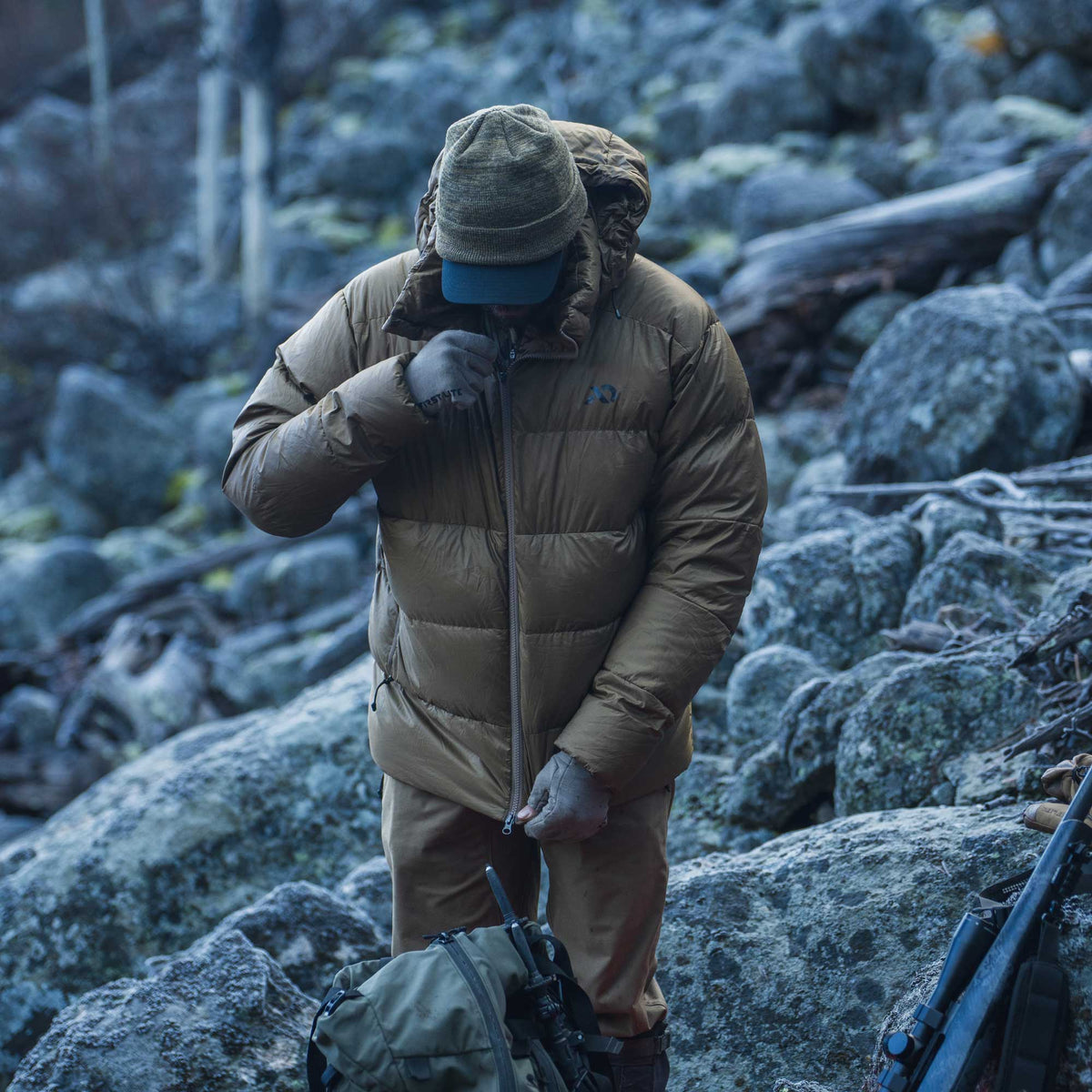 A person standing on a rocky outdoor terrain wearing a tan insulated puffer jacket, matching pants, gloves, and a cap, with a backpack in the foreground.