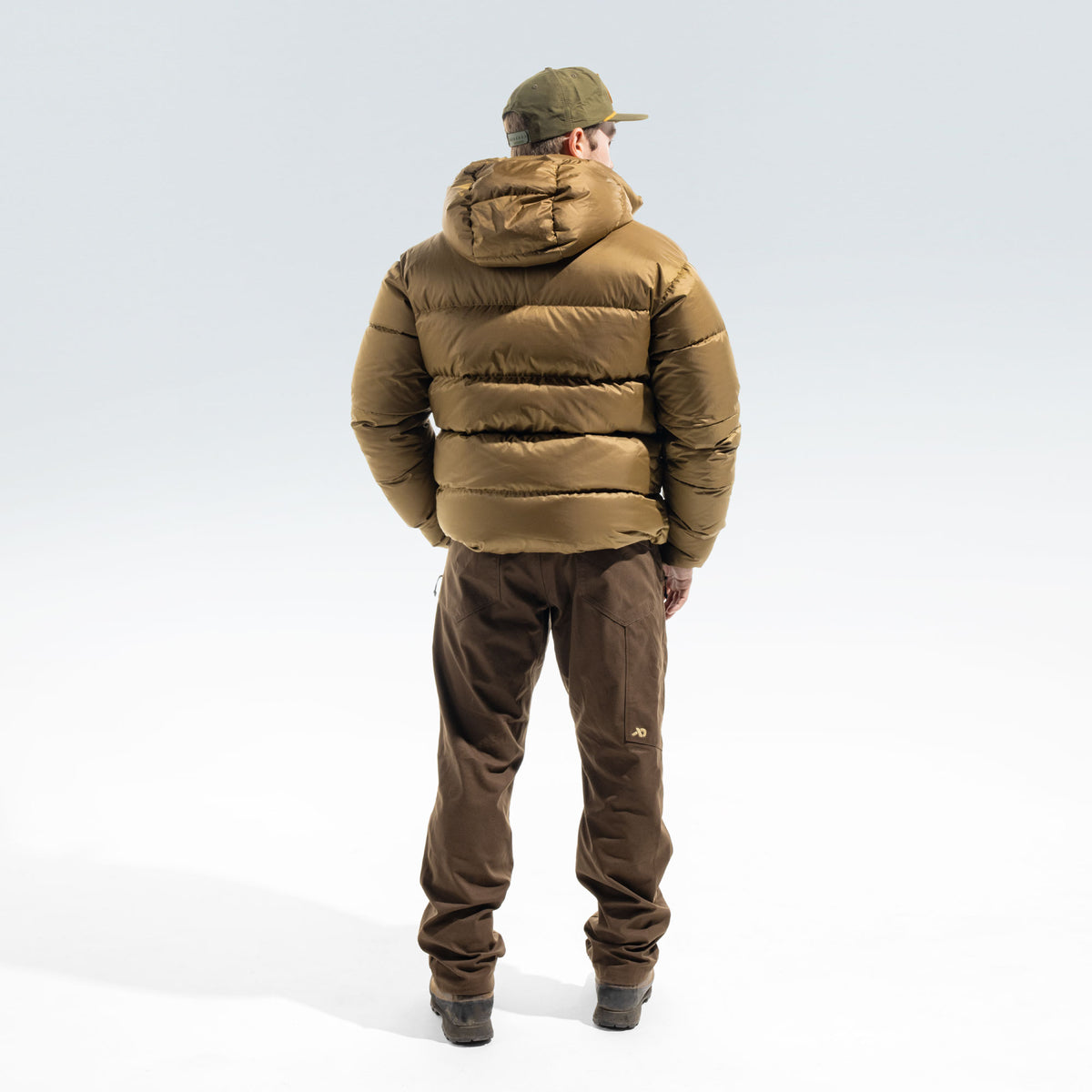 A man viewed from behind wearing a brown puffer jacket, brown pants, and an olive green cap, standing against a light gray studio background.