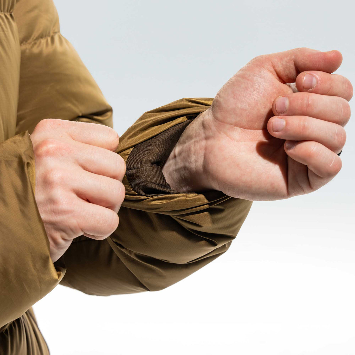 Close-up of a person's hands and arms pulling up the cuff of a brown puffer jacket to reveal a breathable mesh lining.