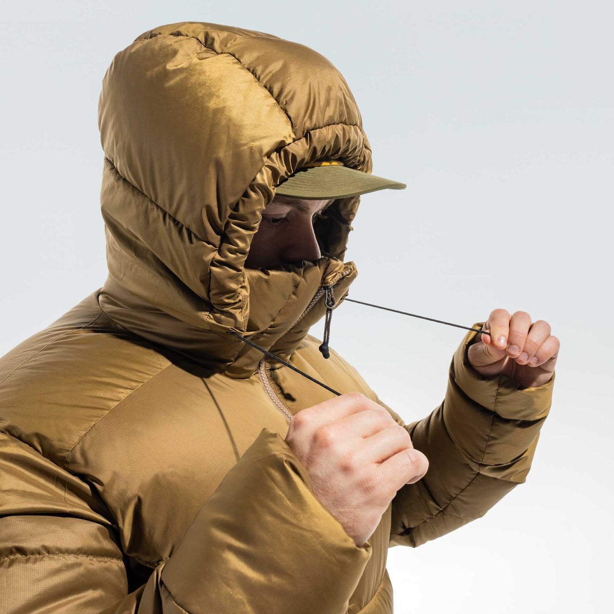 A person wearing a tan hooded puffer jacket and pulling the drawstring of the hood, with a light gray background.