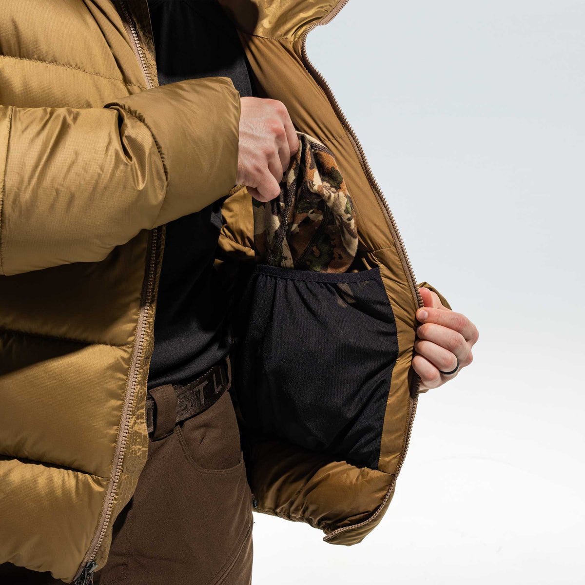 Close-up of a person wearing a camel brown puffer jacket, opening the inner pocket to reveal a camouflage-patterned garment inside, against a clean white background.