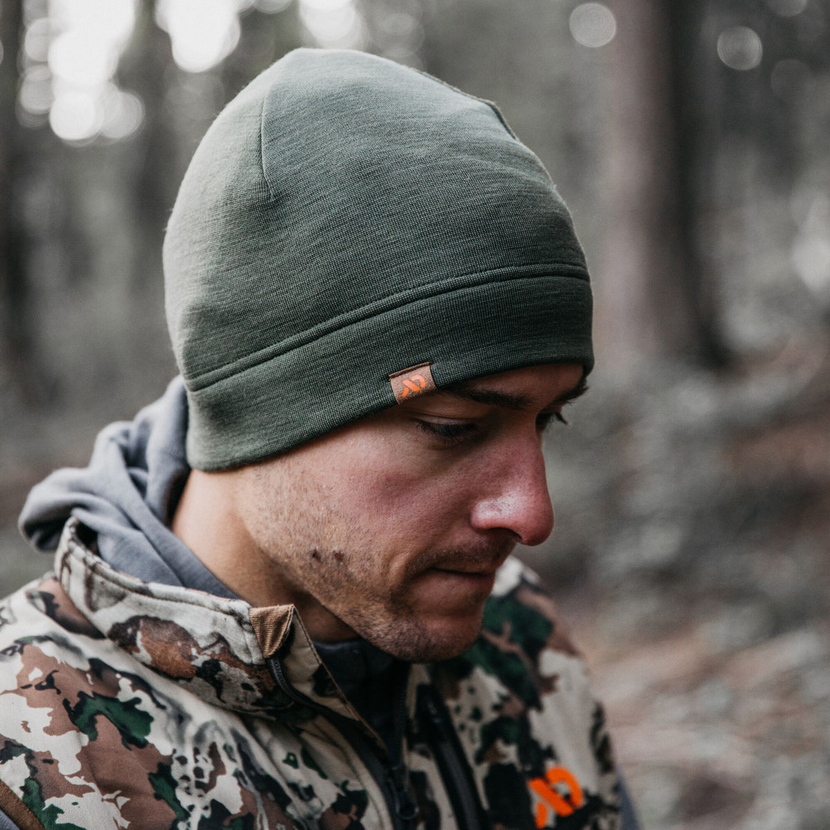Dark olive green knit beanie featuring a small brown patch with an orange logo, worn by a man.