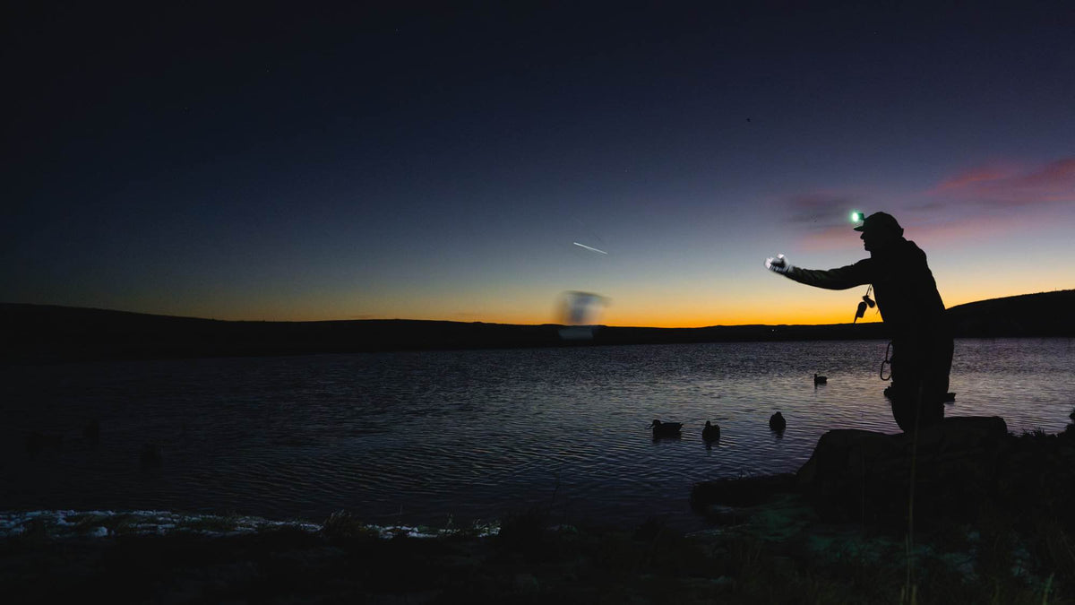 Bright green headlamp illuminates person's silhouette by a dark lake with duck decoys.