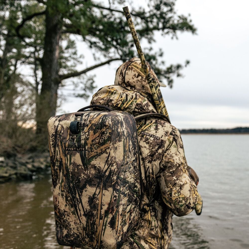 First Lite camouflage dry bag backpack with a top handle and logo, worn on a hunter's back by the water.