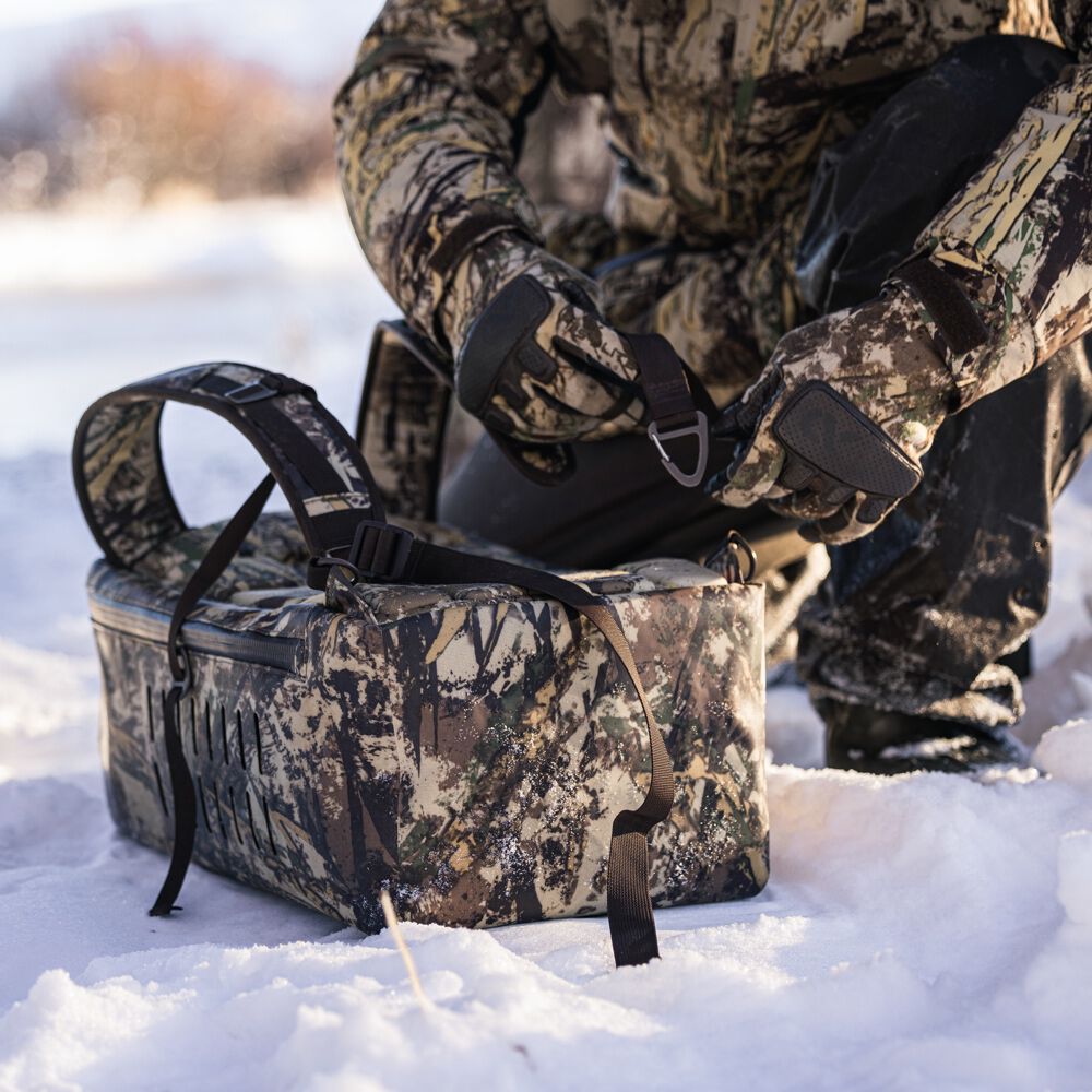 Camouflage soft cooler bag with a top zipper, padded shoulder strap, and side webbing sits in snow as a person secures a strap with a metal D-ring.