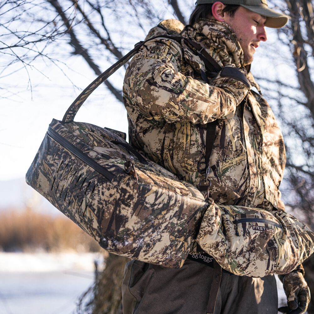Camouflage game bag with a top zipper and side ventilation slits carried by a hunter wearing a matching First Lite hand muff.
