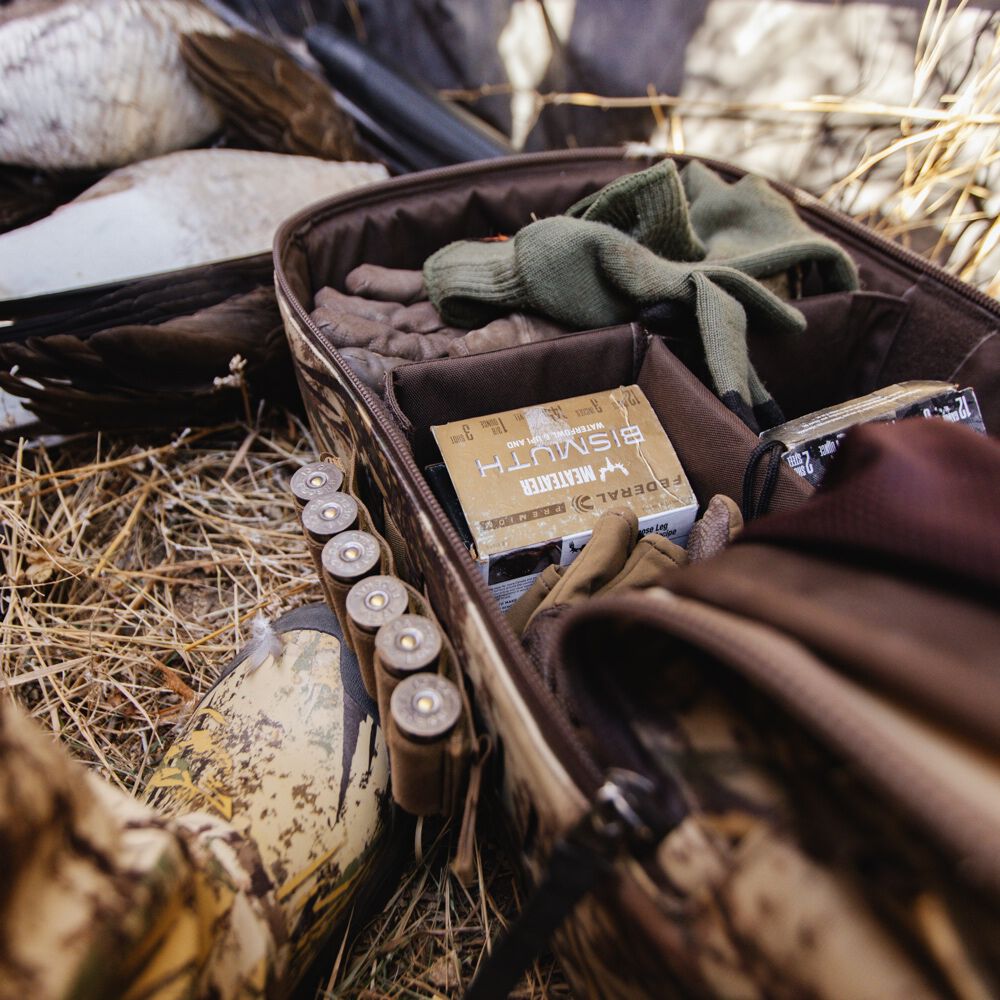 An open camouflage hunting gear bag displays organized compartments holding green gloves, brown gloves, a box of Federal Bismuth shotgun shells, and an external holder with more shells.