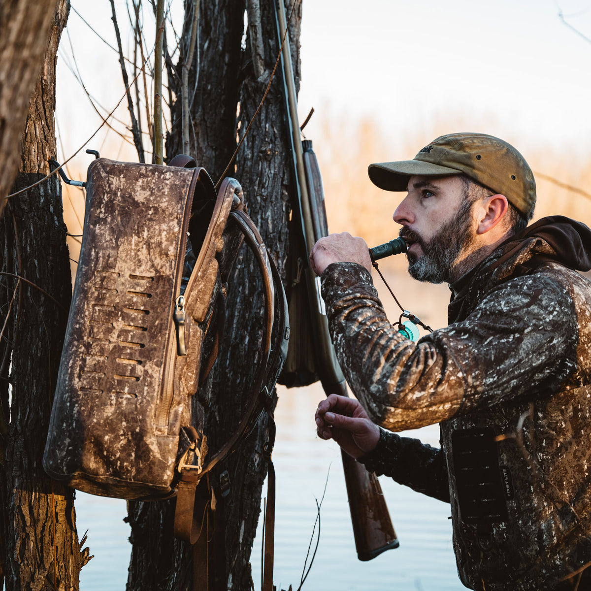 Hunter in camouflage blows a duck call next to a camouflage backpack with slotted vents and a rifle hanging from trees.