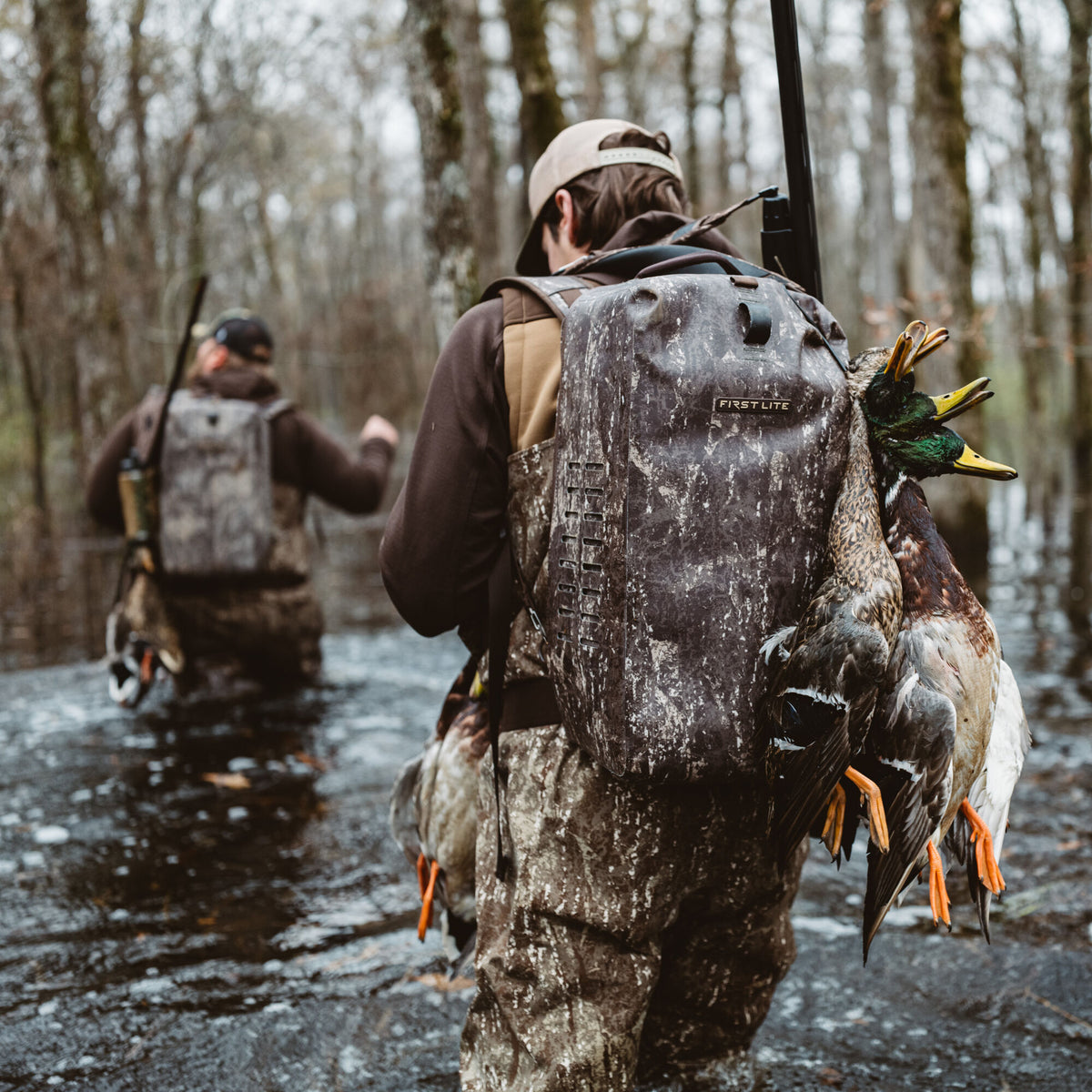 A hunter wears a muddy First Lite dark camouflage backpack with three harvested ducks and a rifle, wading through a forested swamp.