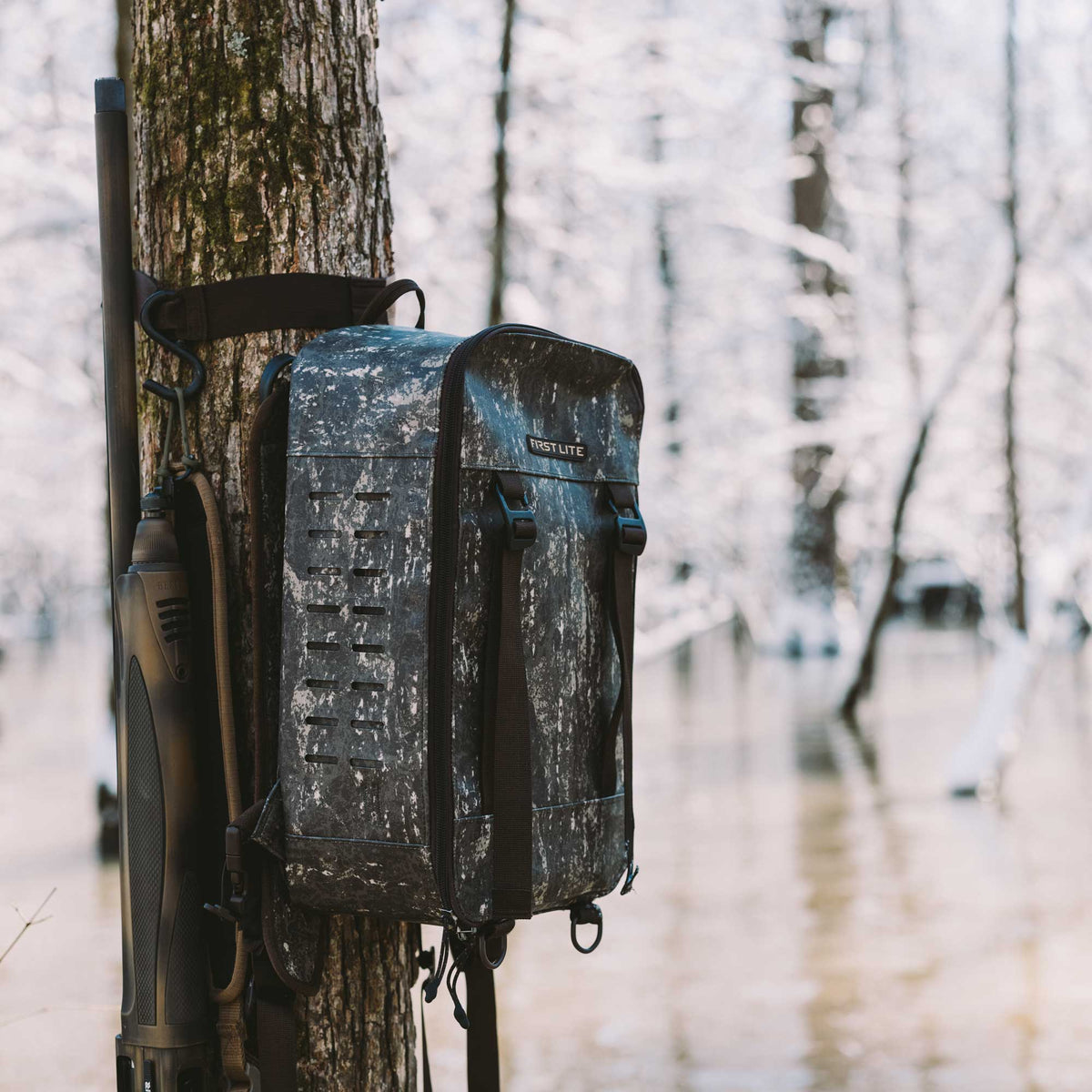 First Lite dark camouflage backpack with horizontal attachment slits hanging on a tree next to a shotgun in a snowy, flooded forest.