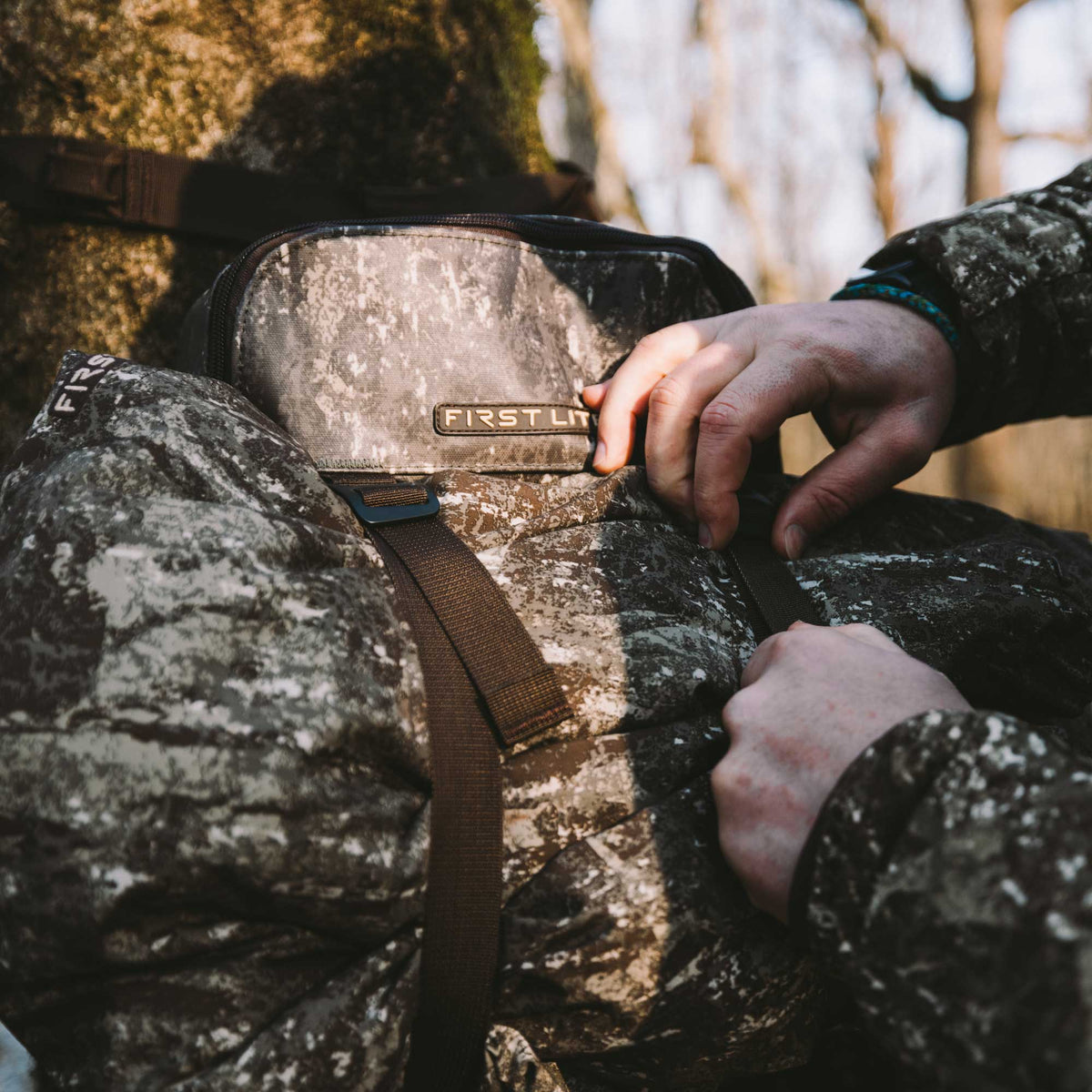 Hands adjust a grey and brown camouflage 'FIRST LITE' gear pouch with a zipper closure and adjustable strap.