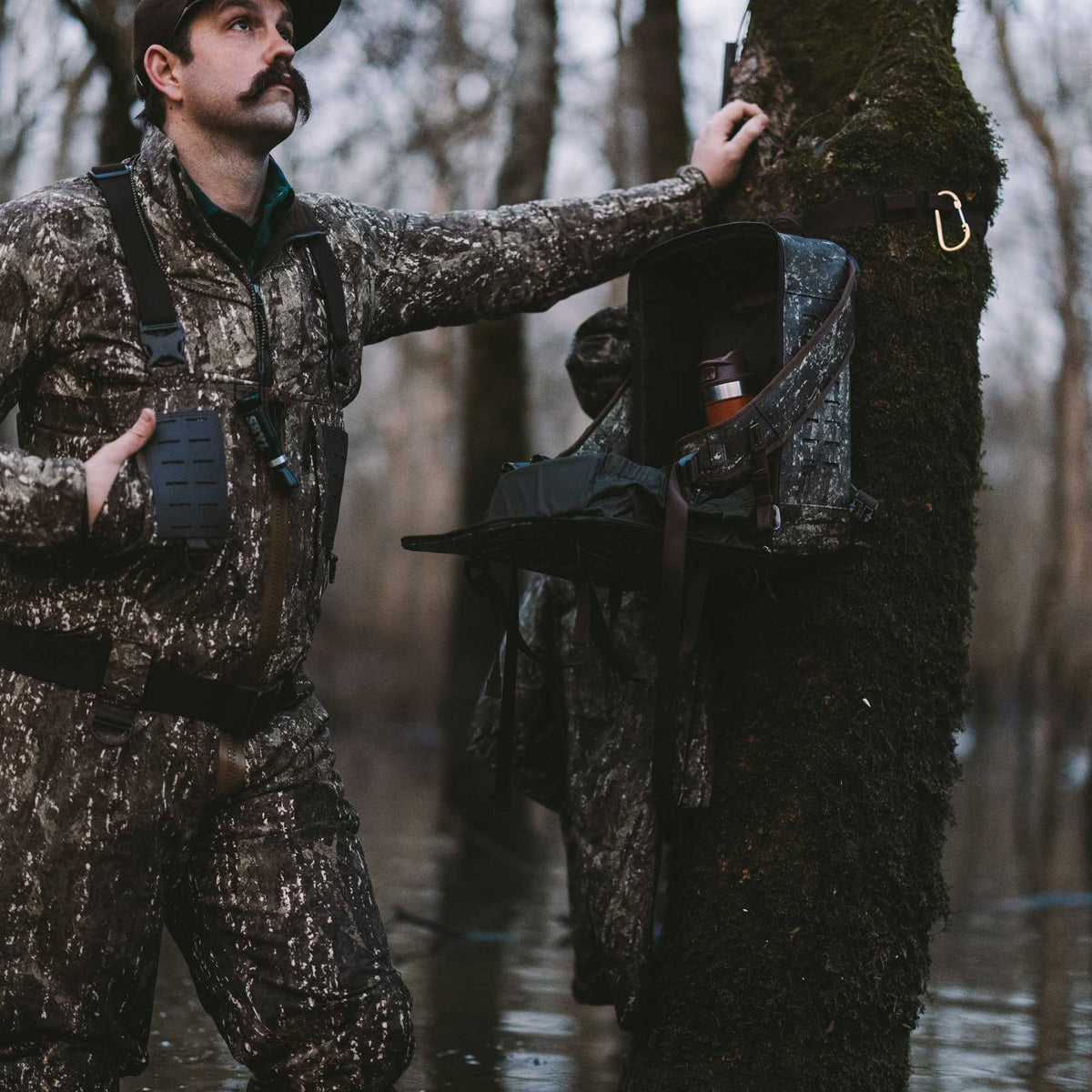 Dark mottled tree-mounted gear box with an open top revealing an orange thermos, secured by black straps and a gold carabiner to a mossy tree.