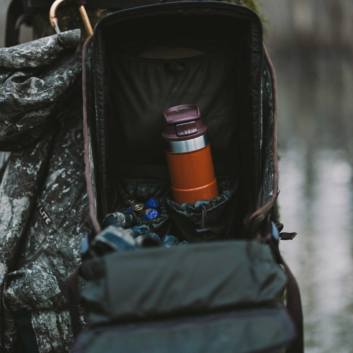 Interior view of a dark open backpack revealing an orange travel mug with a maroon lid and blue shotgun shells, beside a camouflage jacket.