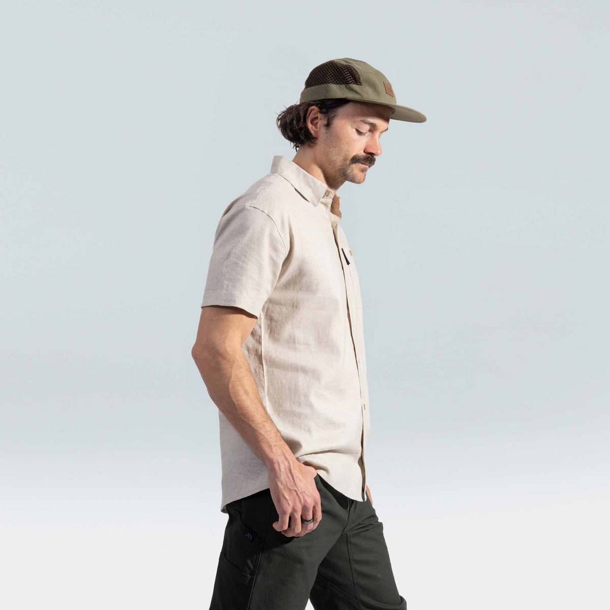 Man wearing a beige shirt, dark pants, and a green cap against a light blue background